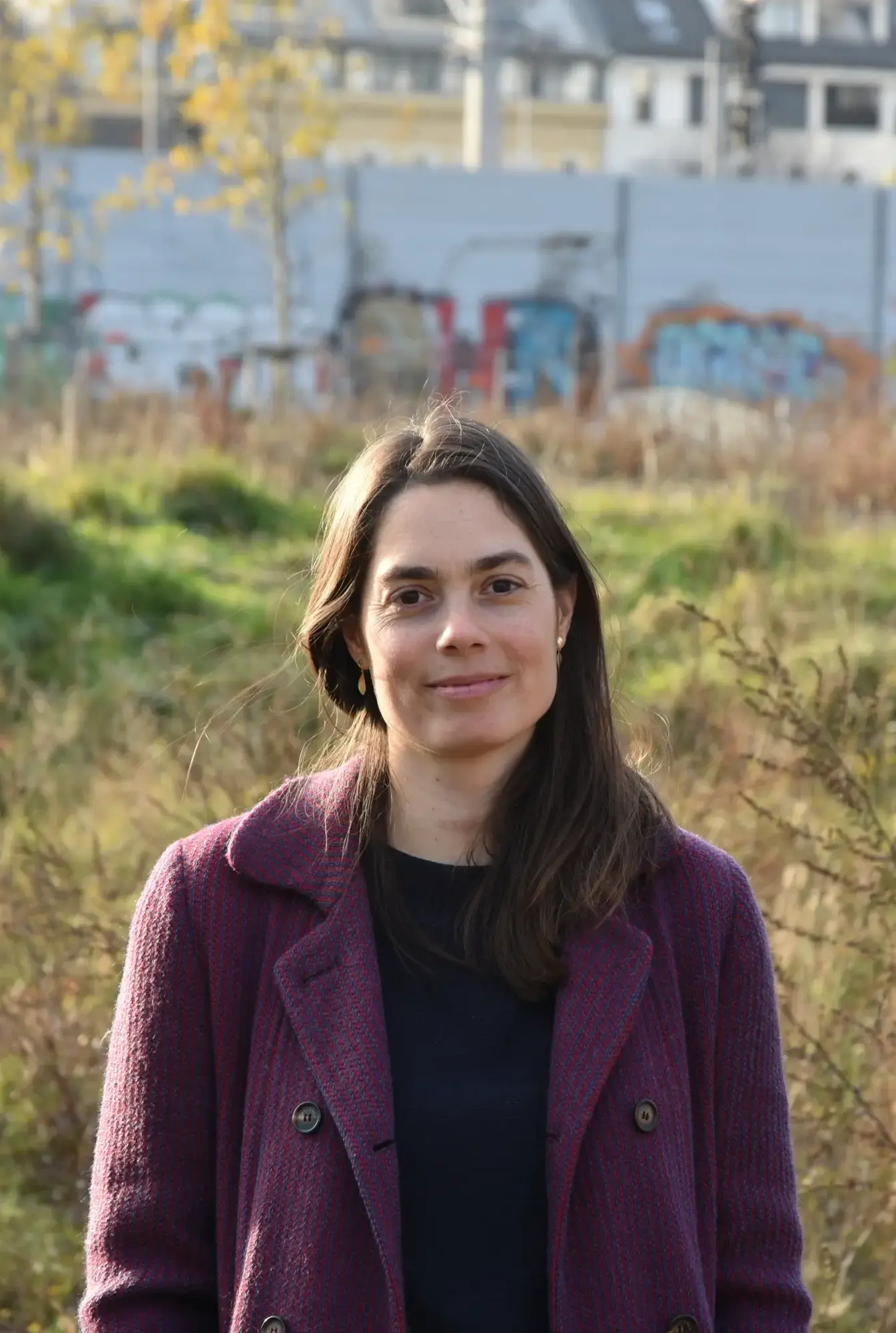 Eine Frau mit langen dunklen Haaren trägt einen dunkelroten Mantel und steht vor einer grünen, mit Gras und Sträuchern bewachsenen Fläche.