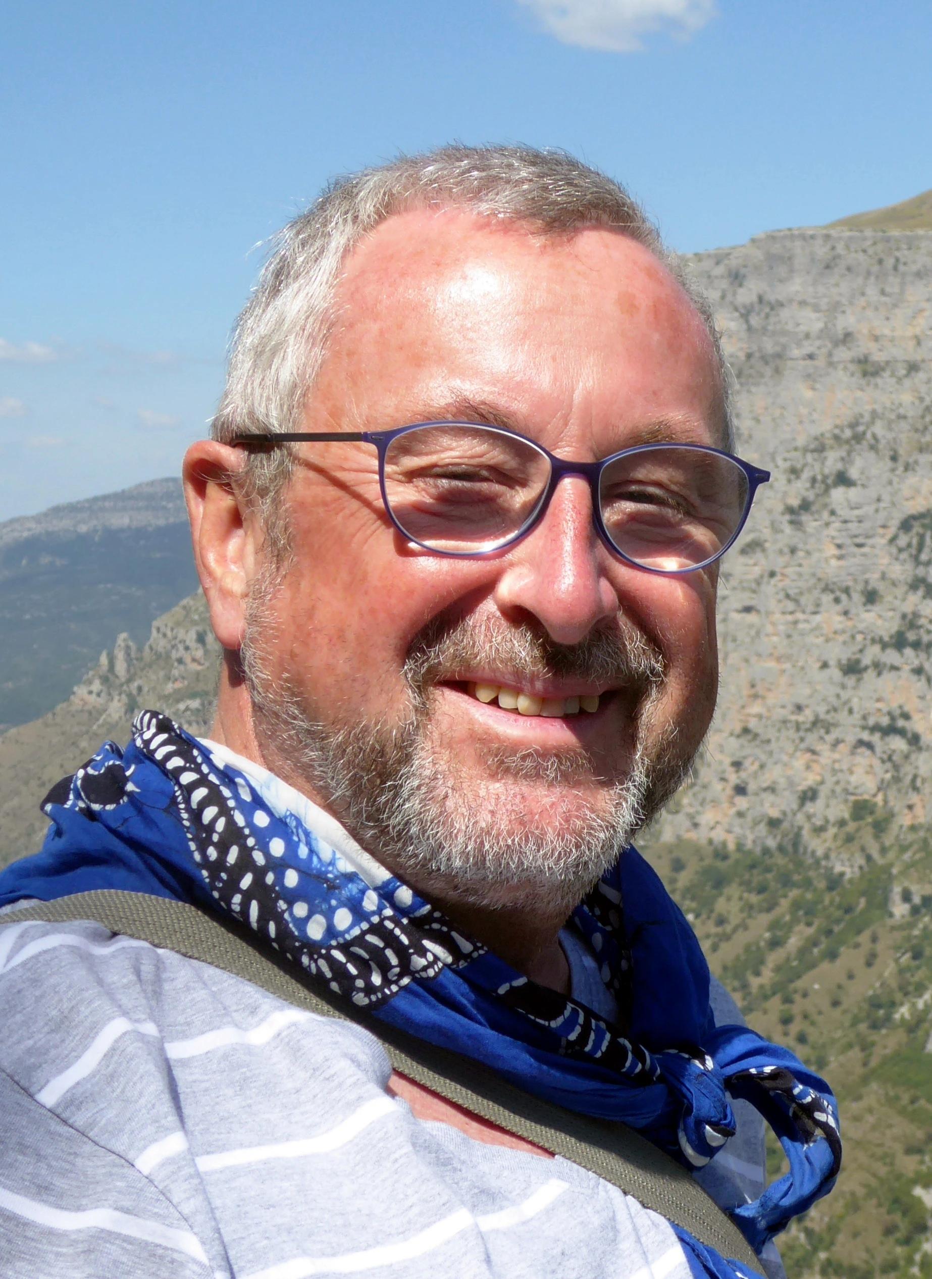 Porträt eines lächelnden Mannes mit Brille, grauem Bart und blauem Halstuch vor Berglandschaft und blauem Himmel.