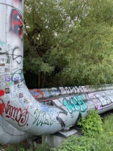 Ein großes, silbernes Rohr mit vielen bunten Graffiti steht auf Betonblöcken vor einem Baum mit grünen Blättern.