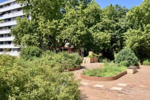 Grün bewachsene Parkanlage mit Bänken, großen Bäumen, Sträuchern und einem mehrstöckigen Wohngebäude im Hintergrund.