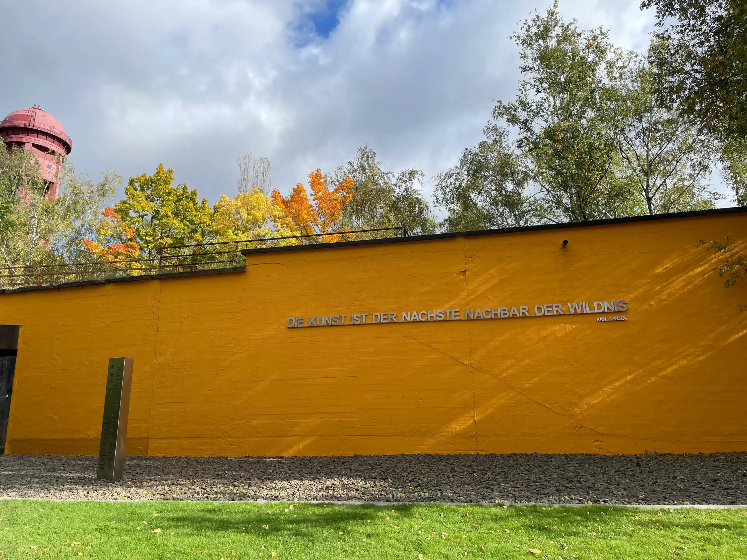 Gelbe Mauer mit dem Zitat Die Kunst ist der nächste Nachbar der Wildnis von Karl Ganser, davor ein grüner Rasen und bunte Bäume im Hintergrund.