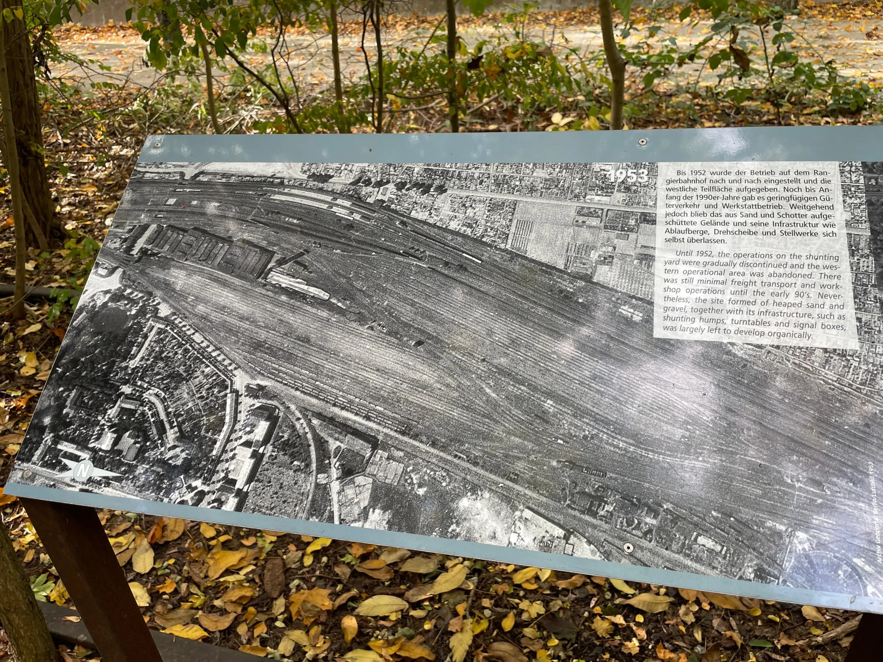 Infotafel mit Luftbild von 1953 zeigt ein großes Bahngelände mit Gleisen und Gebäuden, umgeben von herbstlichem Laub und Bäumen.